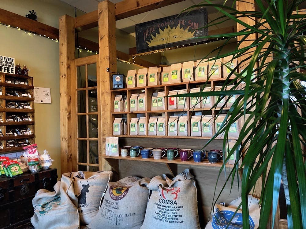 The shelf wall at the Lakewood shop, stacked with bags of coffee and mugs under a folk art sun sign
